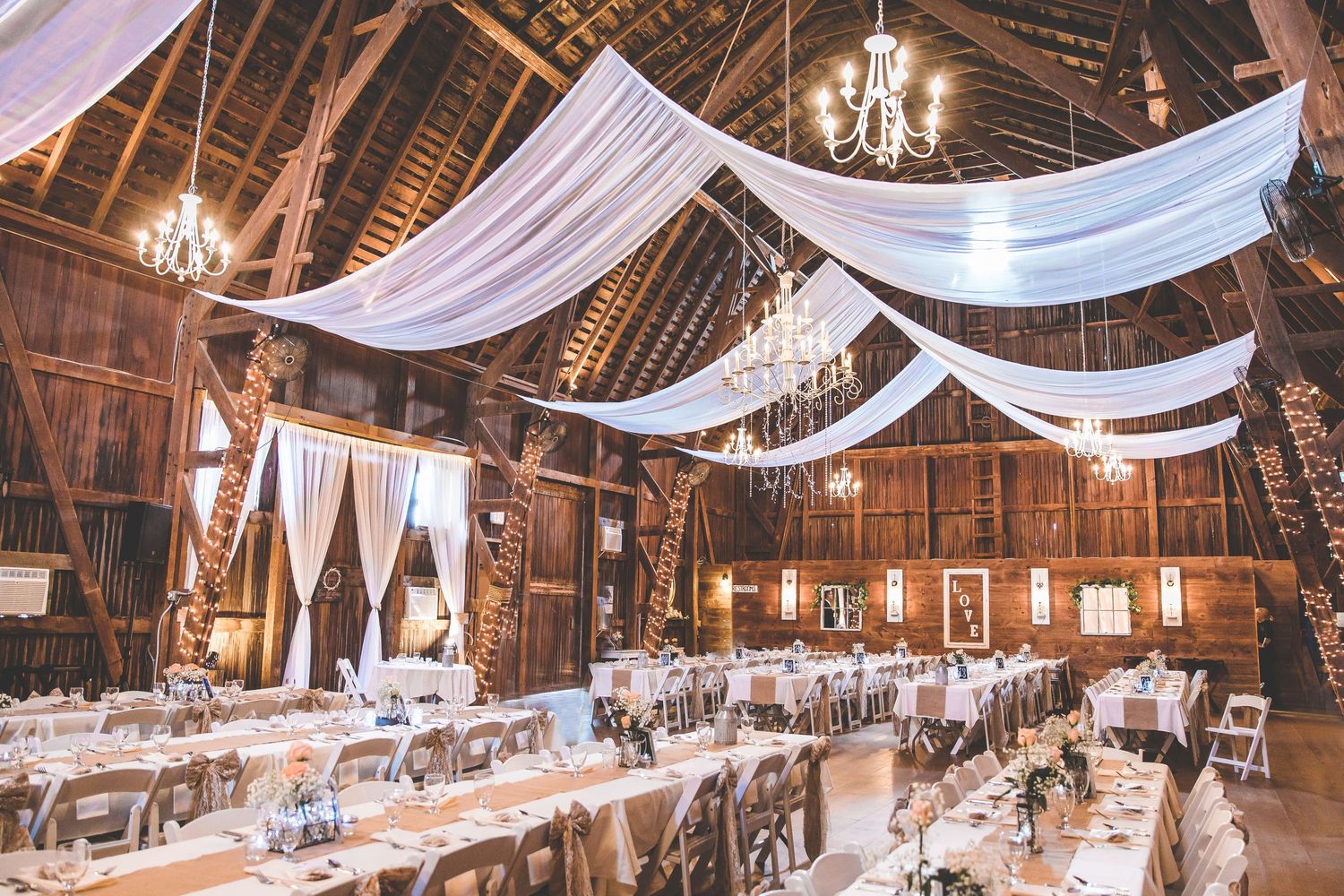 Rustic barn wedding reception venue with elegant white draping and chandeliers.