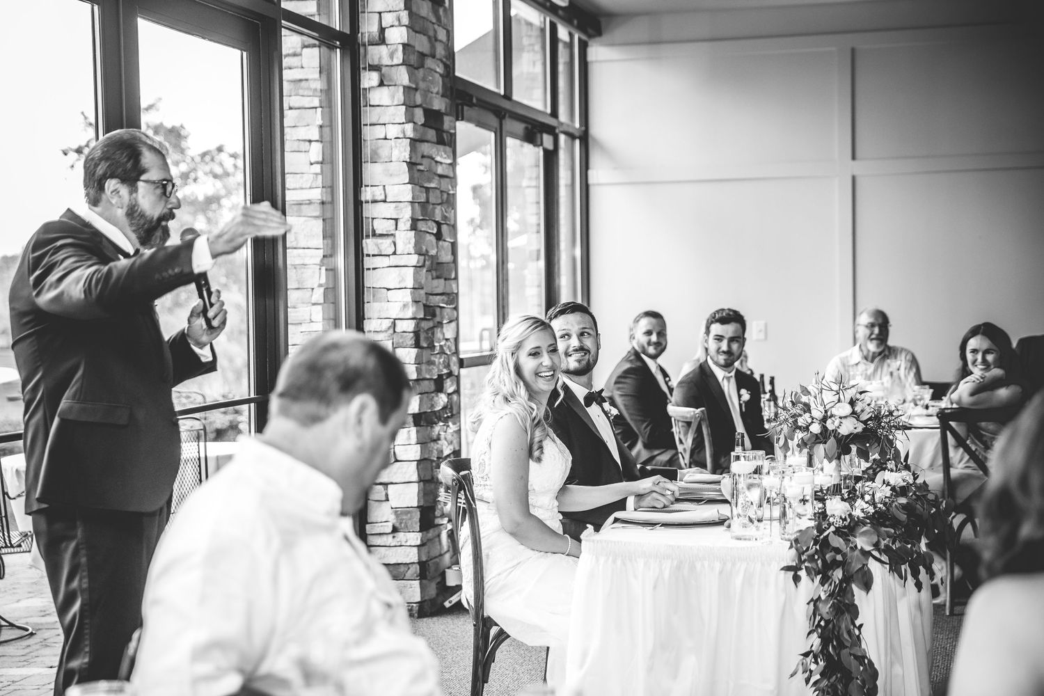A wedding toast being given during a reception while guests listen at decorated tables.