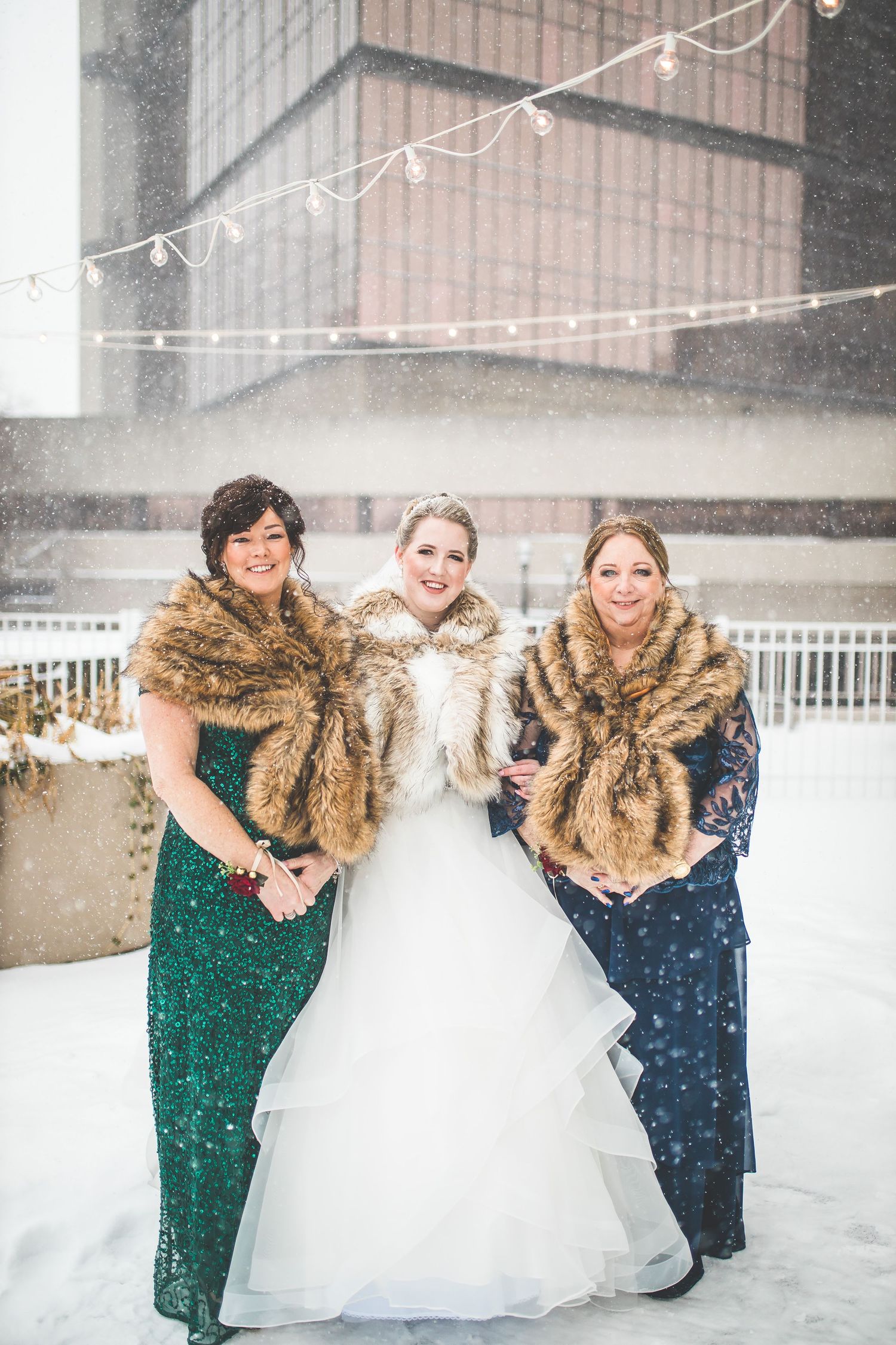 Three women in formal attire and fur stoles pose together in snowy winter setting with string lights.