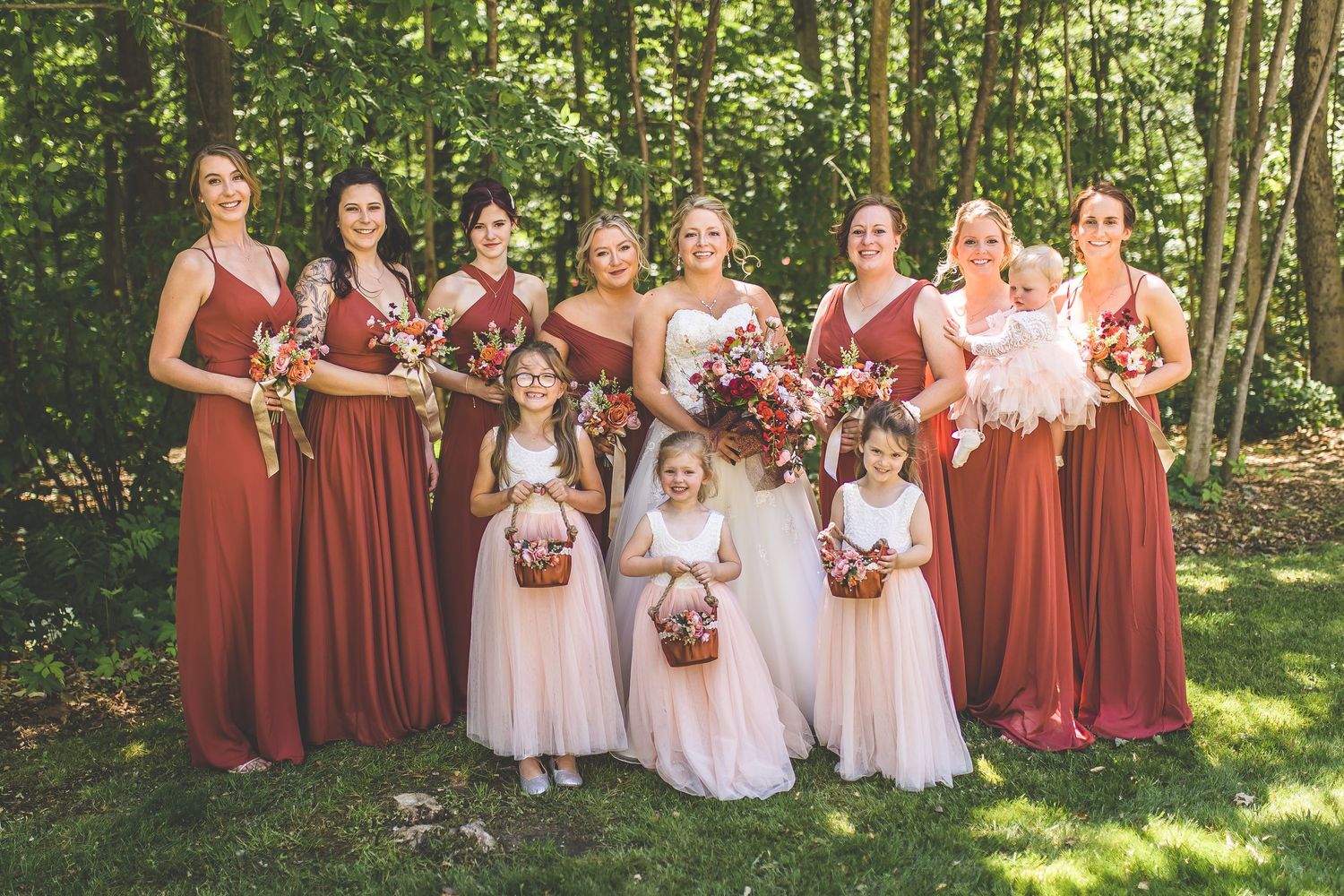 Wedding party poses in burgundy and white dresses against a lush forest backdrop.