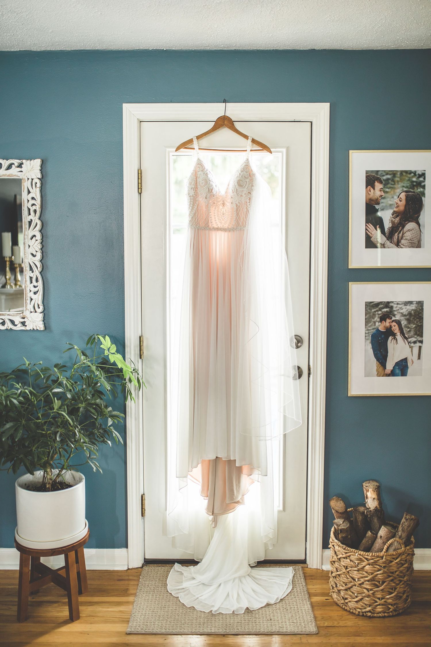 A blush wedding dress hangs elegantly on a door against a teal wall with framed photos and potted plant.