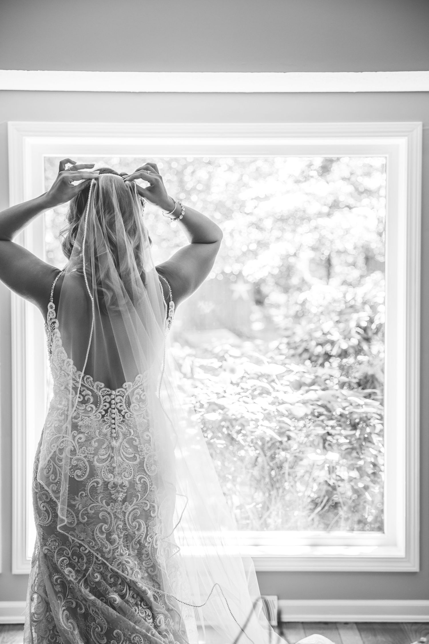 A back view of a bride adjusting her veil by a window in a lace detailed wedding dress.