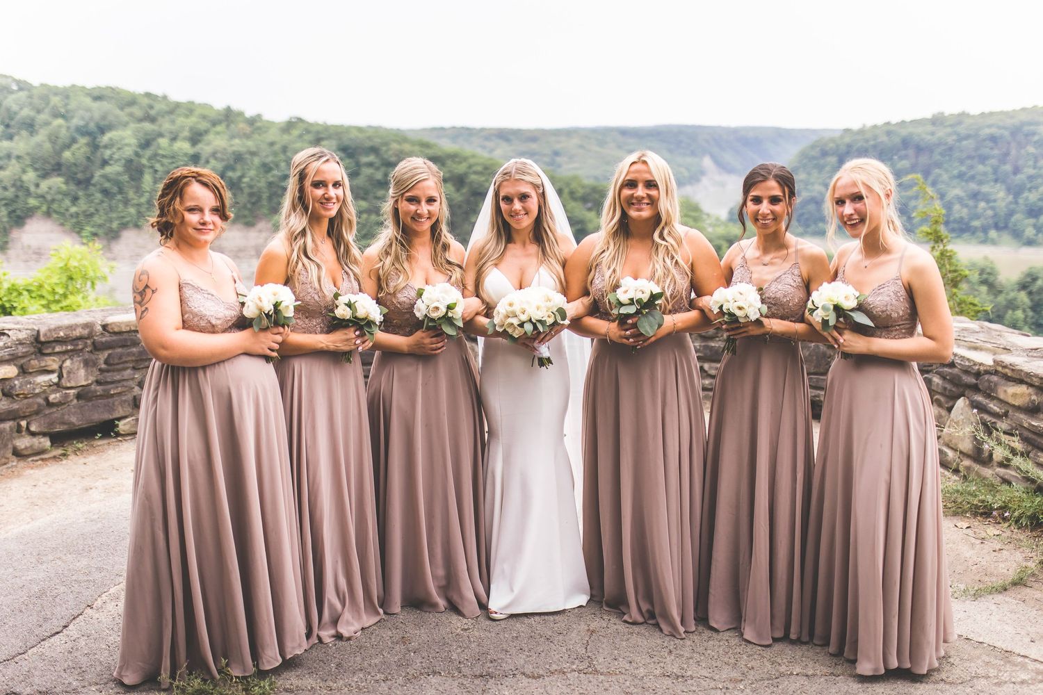 Bridesmaids in flowing mauve dresses pose with bride in white against mountain backdrop.