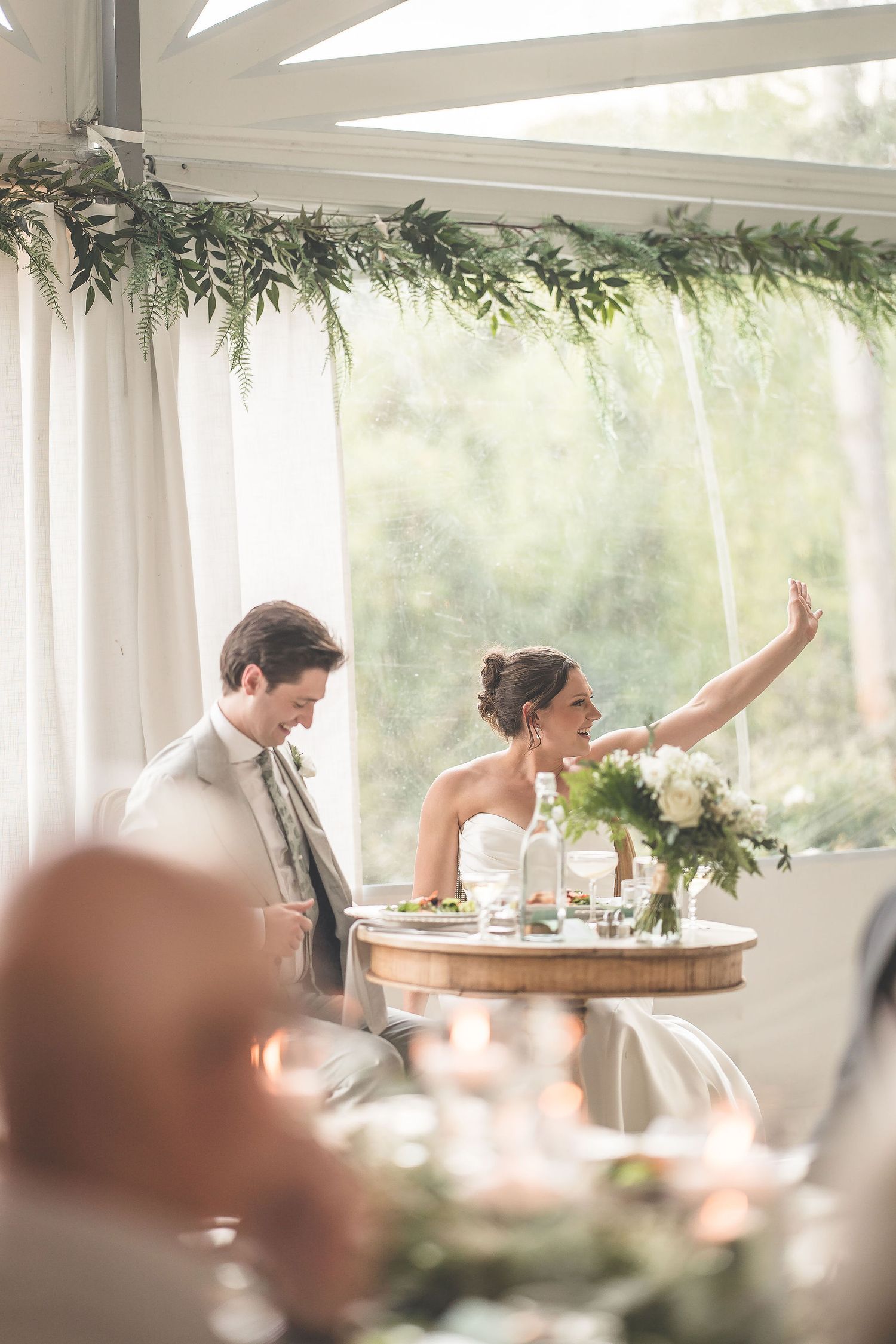 Elegant wedding reception table settings with greenery decor in a bright sunlit tent.