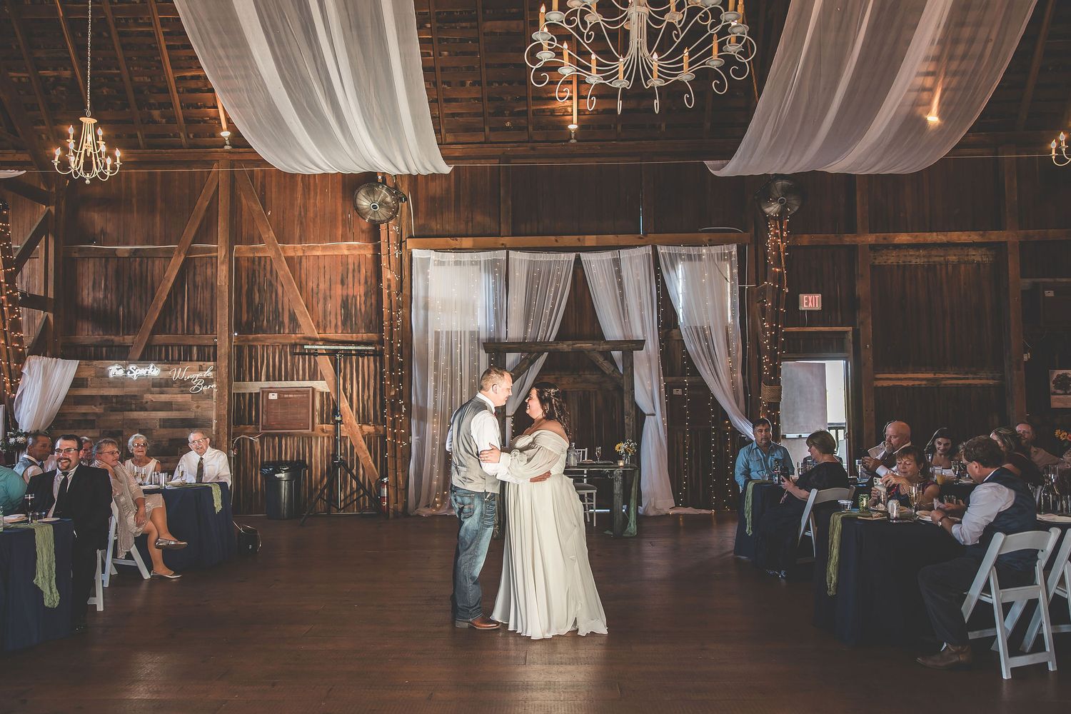Panoramic view of a barn wedding reception with elegant chandeliers and white fabric decorations.