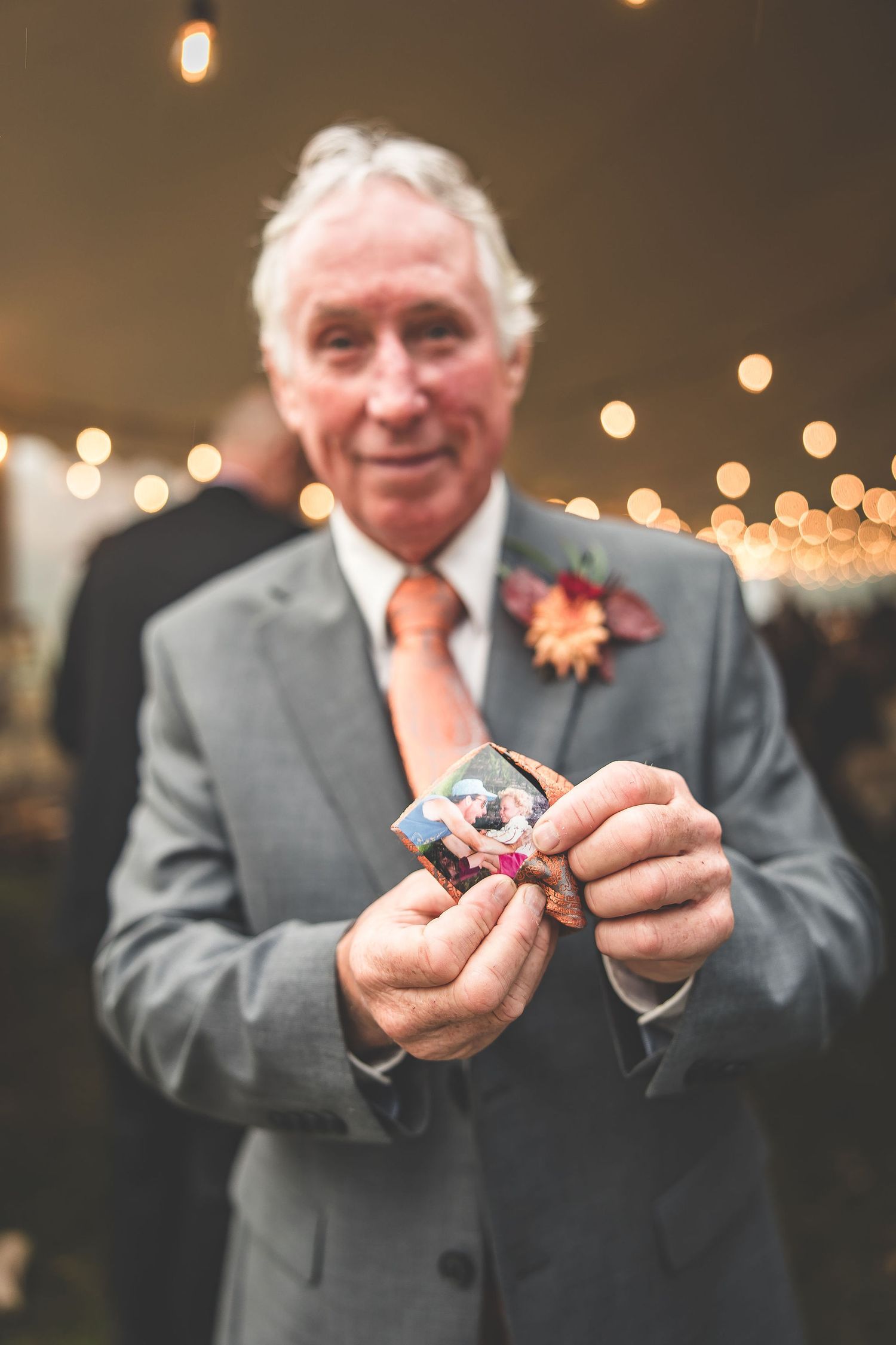 Senior man in grey suit holds small items while smiling under warm lighting at wedding reception.