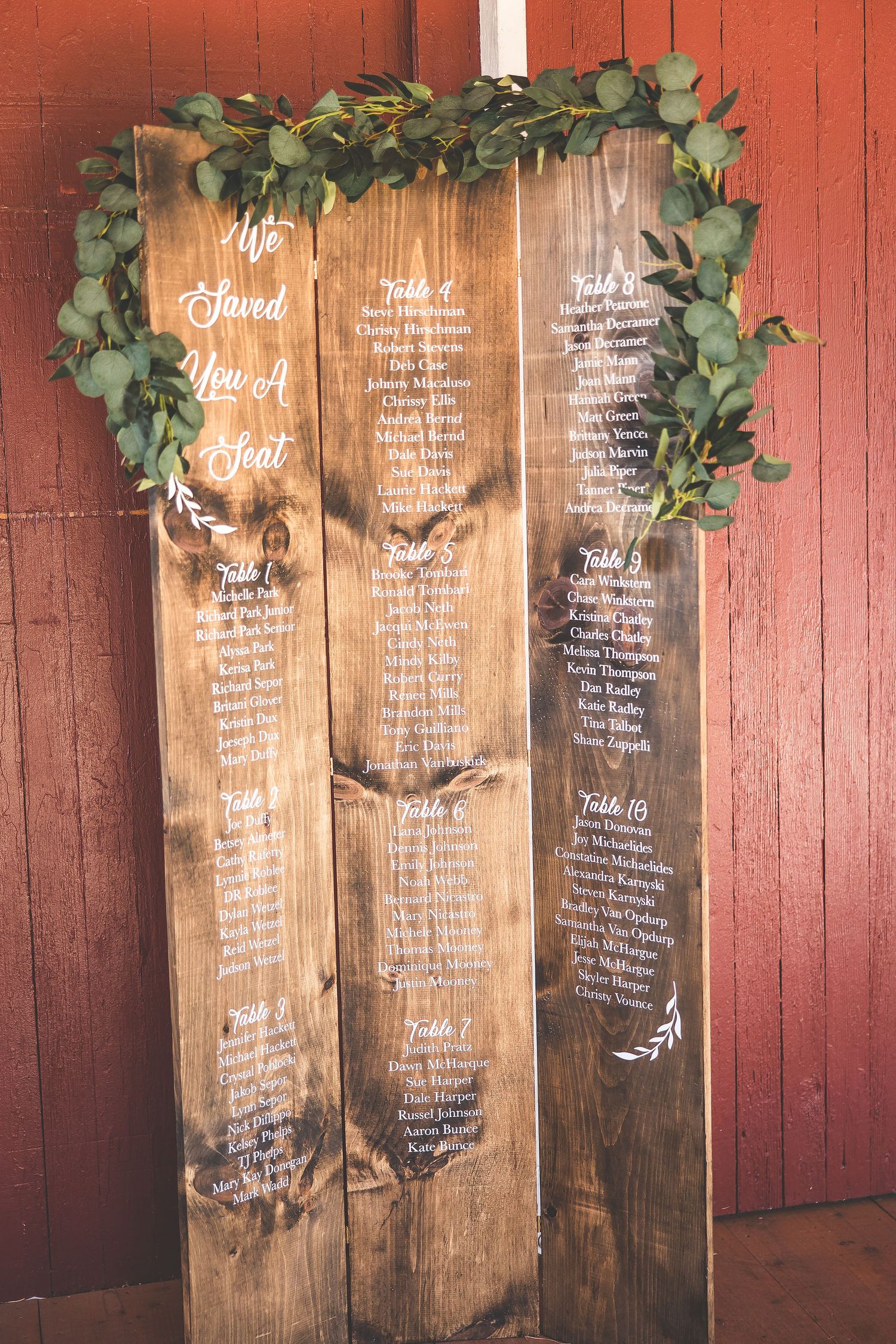 Wooden seating chart boards adorned with greenery against a red barn wall.