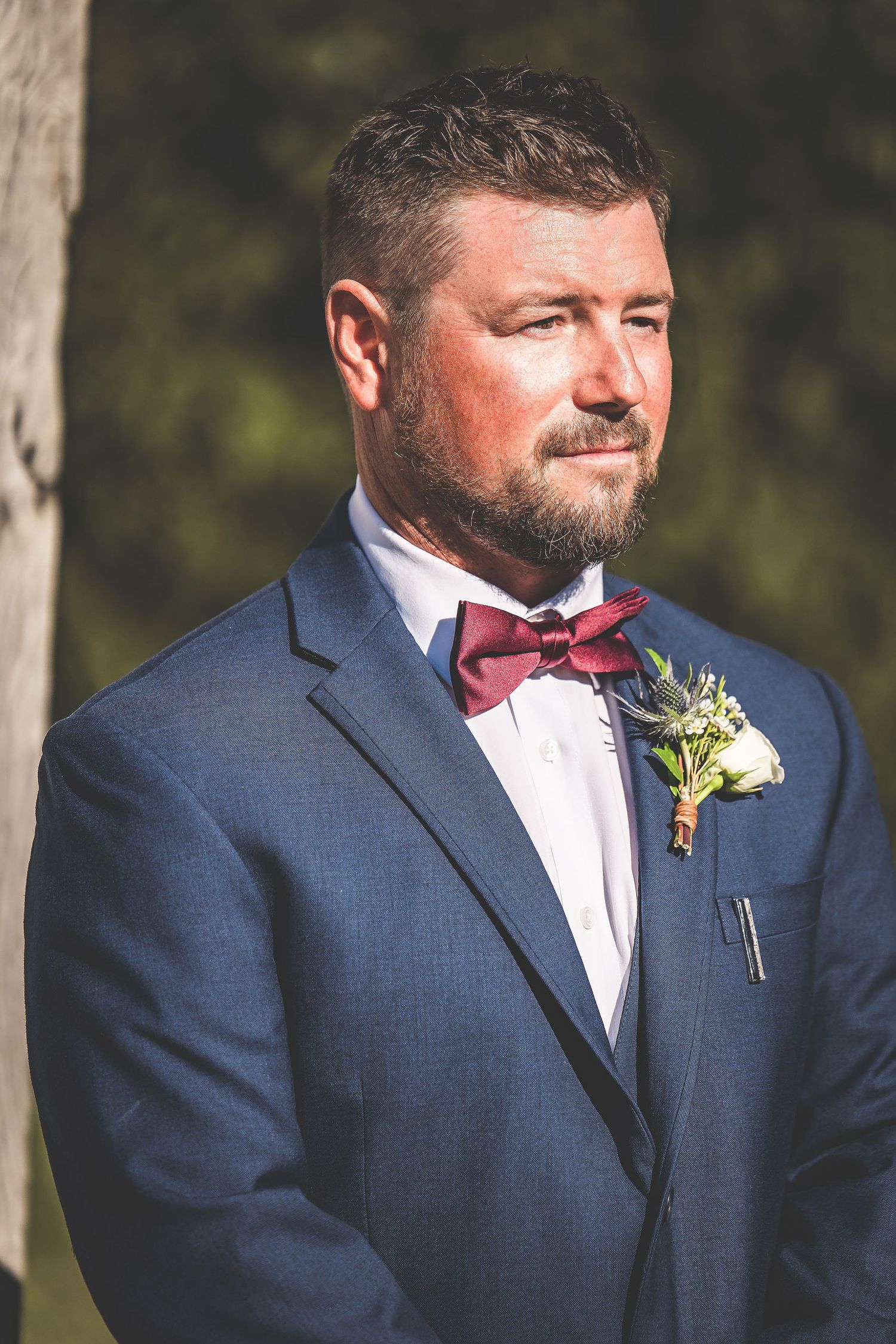Groom in navy suit with boutonniere smiles during outdoor wedding ceremony.