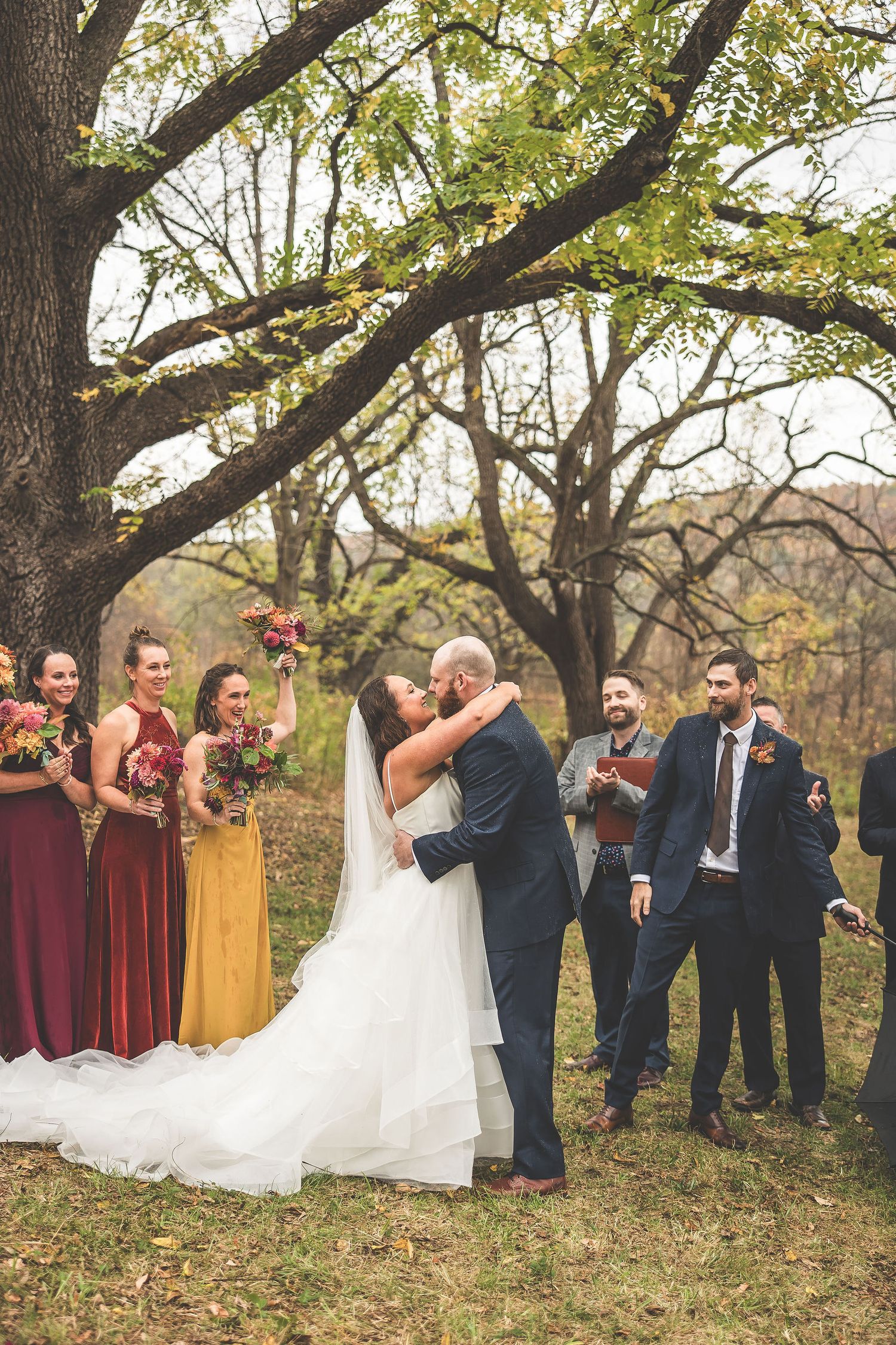 Wedding party celebrates as couple shares first kiss under autumn trees.