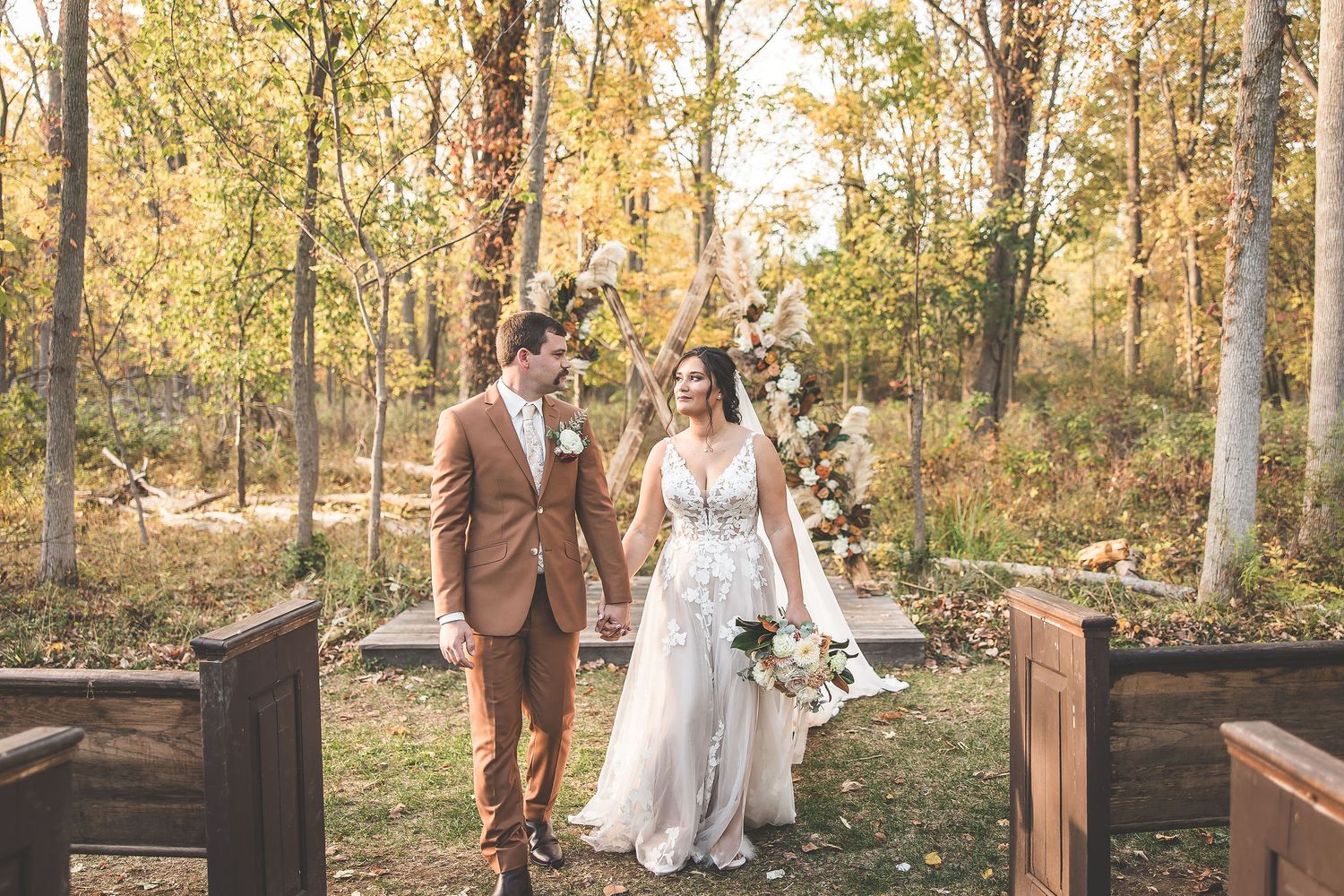 Wedding couple strolls hand in hand along a rustic wooden fence in an autumn forest setting.
