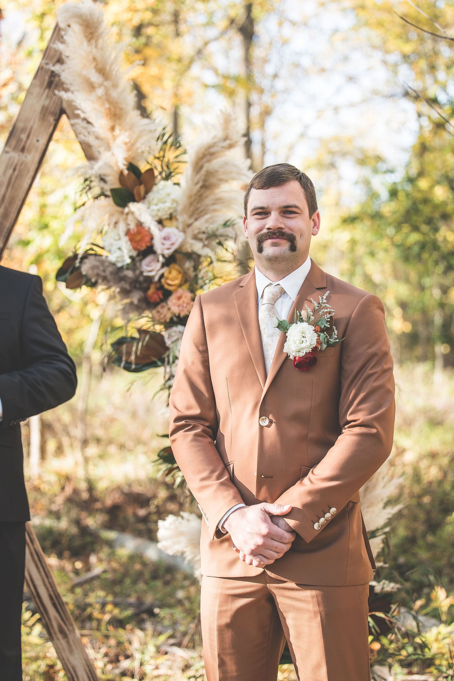 Groom in tan suit stands at rustic wooden arch decorated with fall flowers and pampas grass.