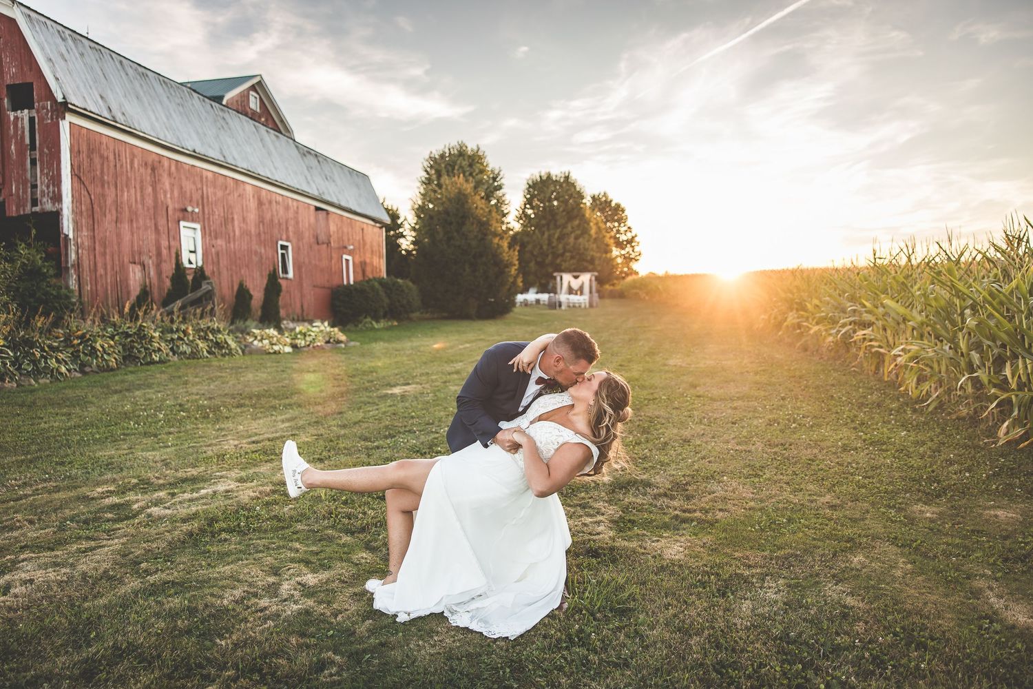 Wedding couple poses dramatically at sunset near a red barn and cornfield.