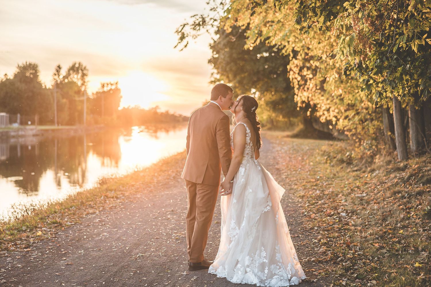 Sunset wedding portraits captured on a scenic path beside water with autumn foliage.