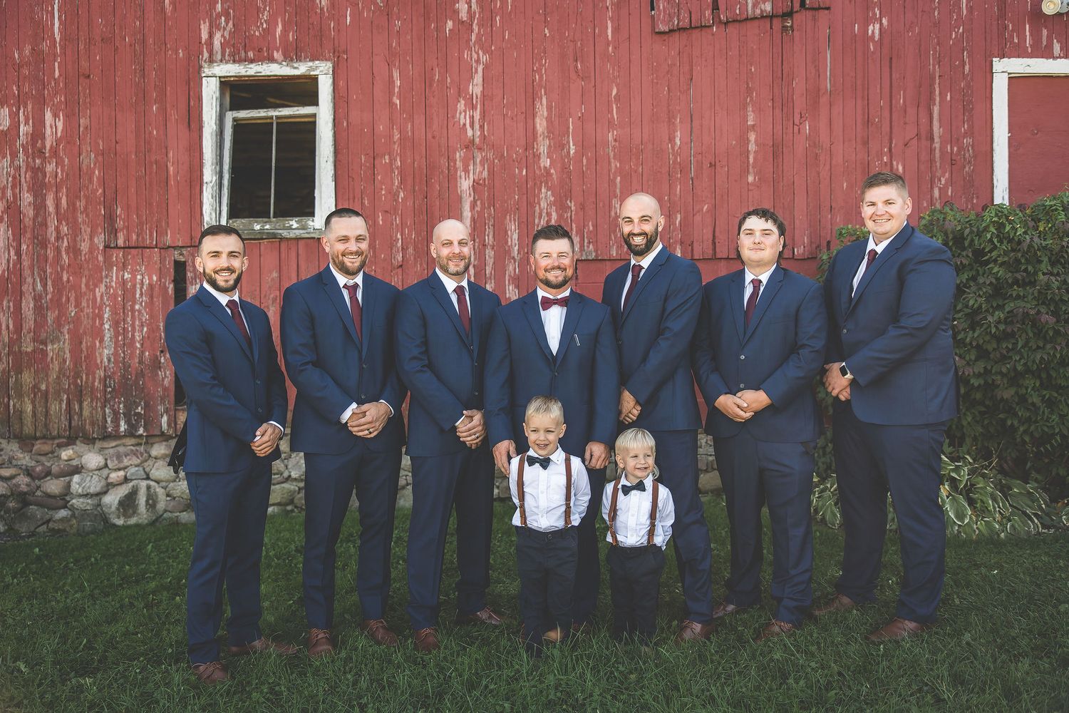 Wedding party poses in formal attire against rustic red barn exterior.