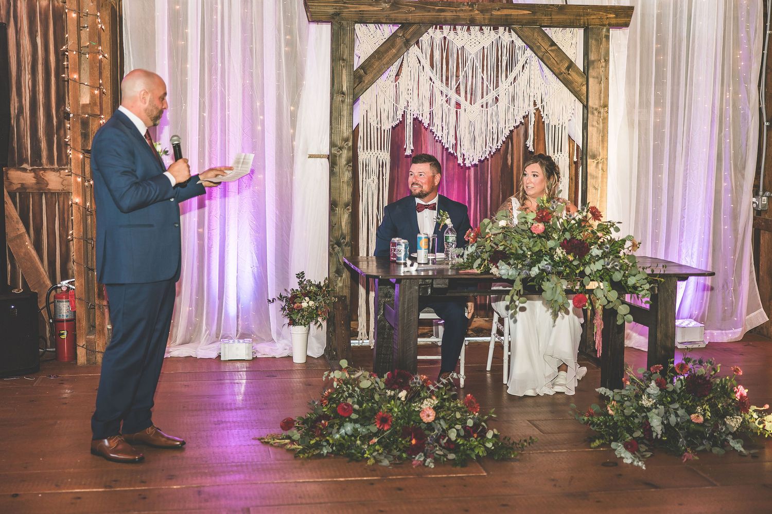 Elegant wedding reception setup with macrame backdrop, wooden beams, and floral arrangements.