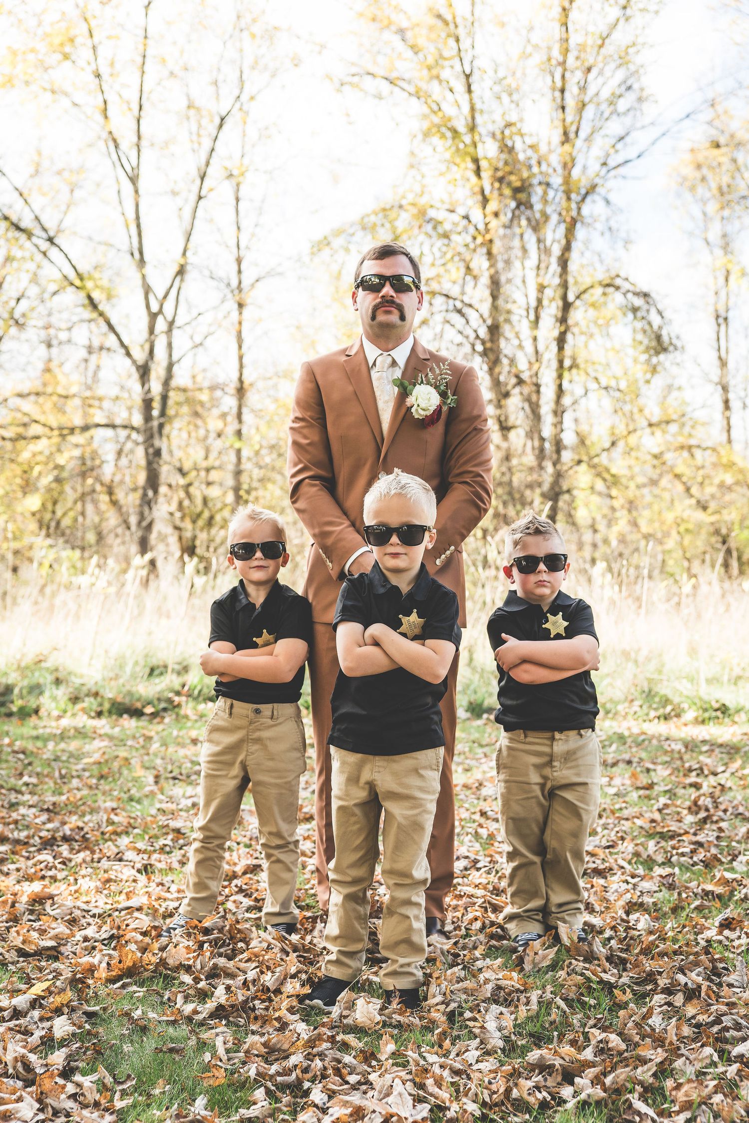 Three young boys in sunglasses pose with an adult in a brown suit for a fun wedding photo.