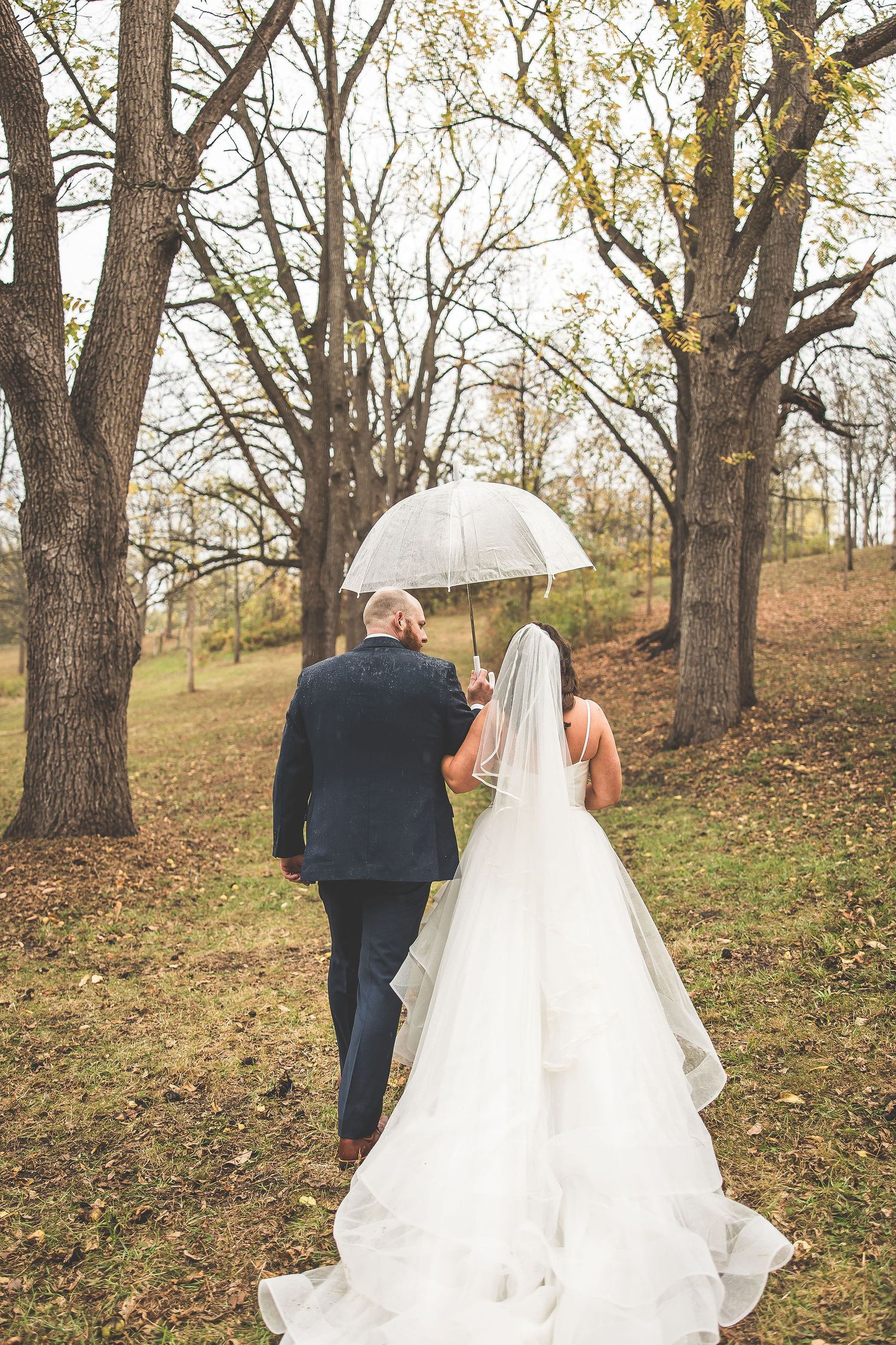 Bride in flowing white gown walks with partner through leaf-covered autumn woodland.