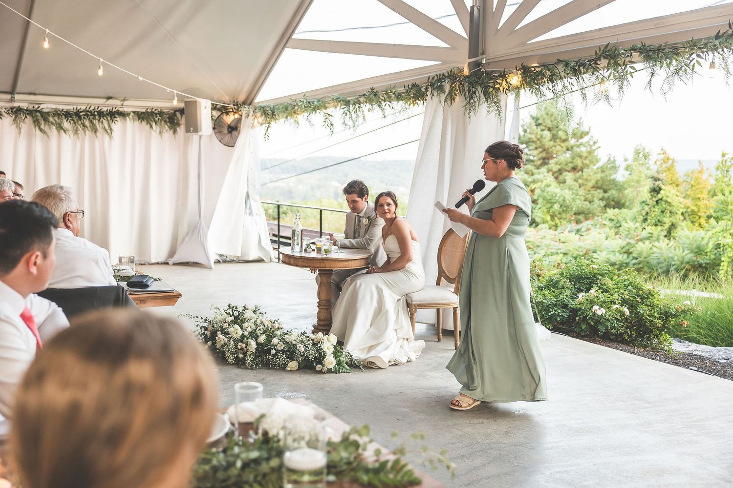 Wedding reception under a white tent with guests watching speeches.