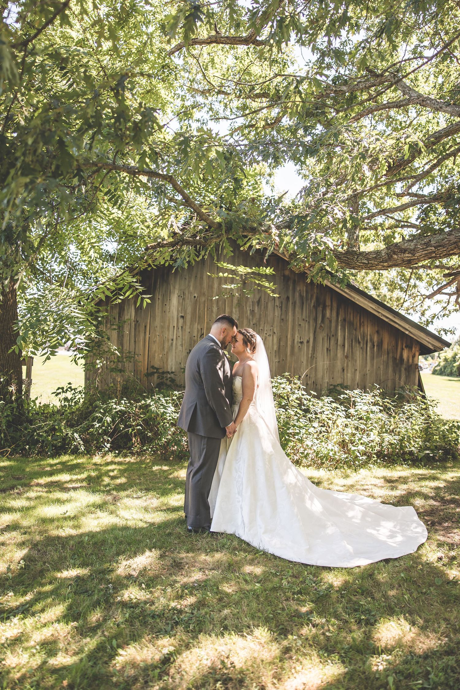 A romantic wedding photo of a couple standing by a rustic wooden barn under tree branches.