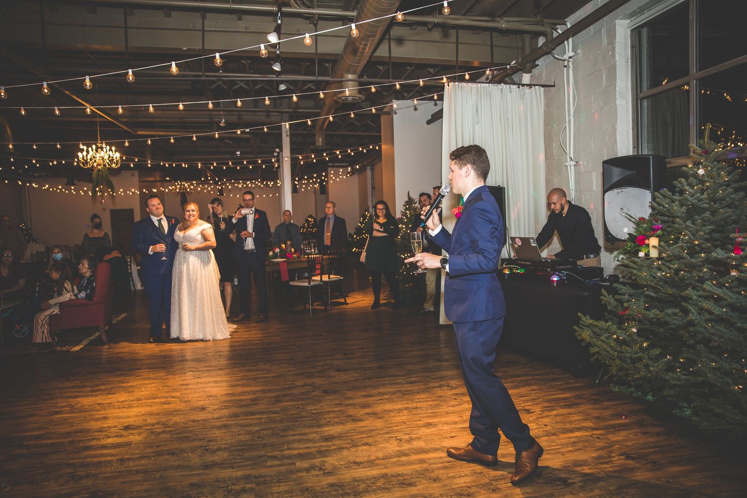 Wedding guests dance and celebrate on a wooden floor under romantic string lights and chandeliers.