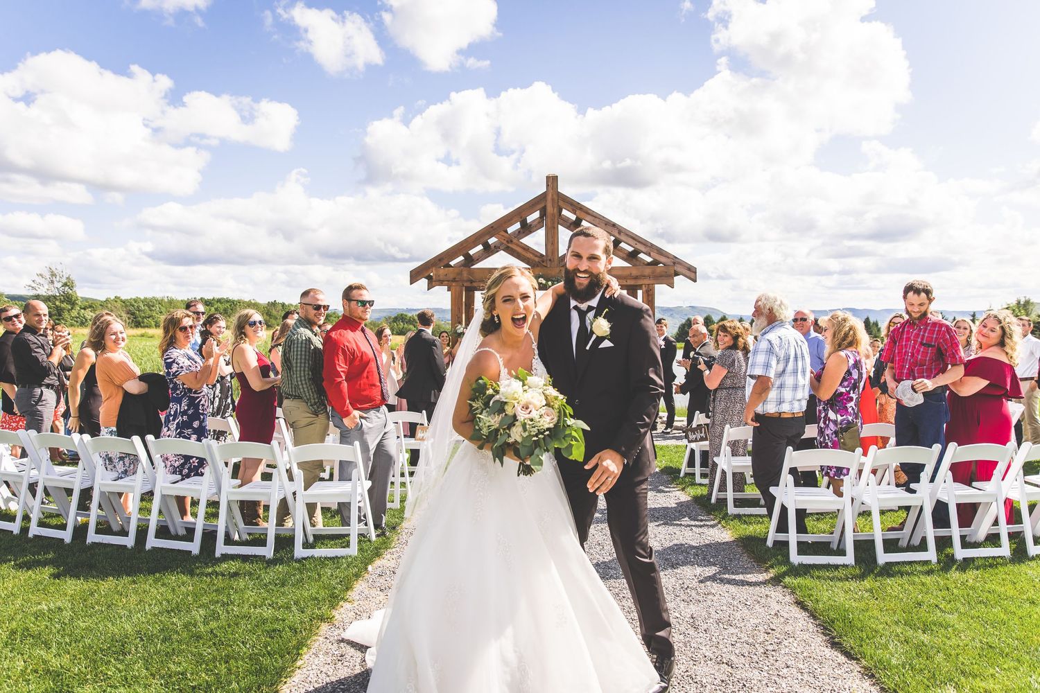 A wedding ceremony processional on a gravel path under a wooden pavilion with white chairs.