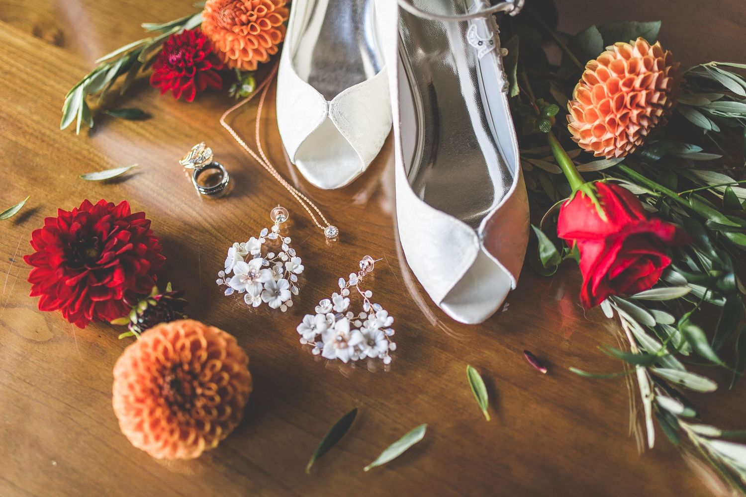 Wedding detail flatlay with orange dahlias, red roses, and bridal accessories on wooden surface.