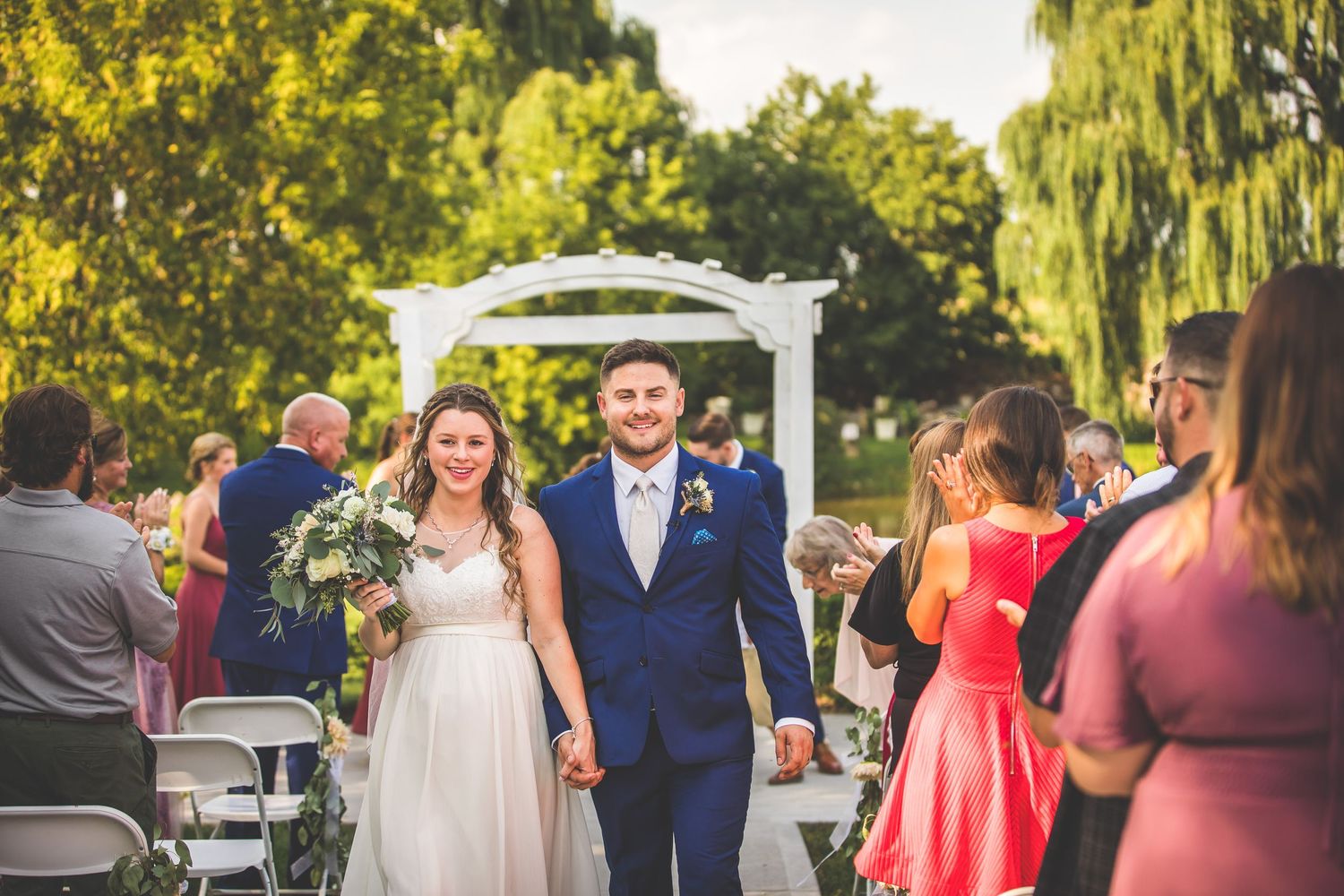 Wedding guests gather on a sunny lawn with bridesmaids in coral dresses and groomsmen in navy suits.