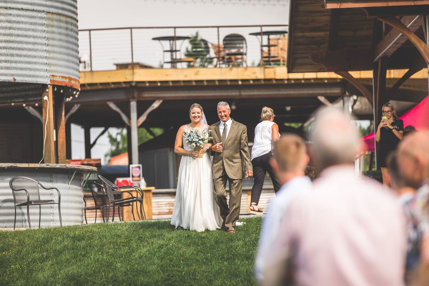 Wedding ceremony guests seated in white chairs on a grassy field with rustic wooden beams and string lights overhead.