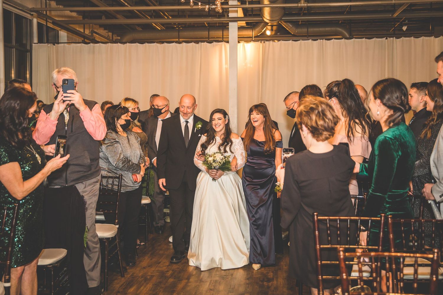 Wedding guests dance and celebrate at an indoor reception venue with warm lighting and rustic decor.