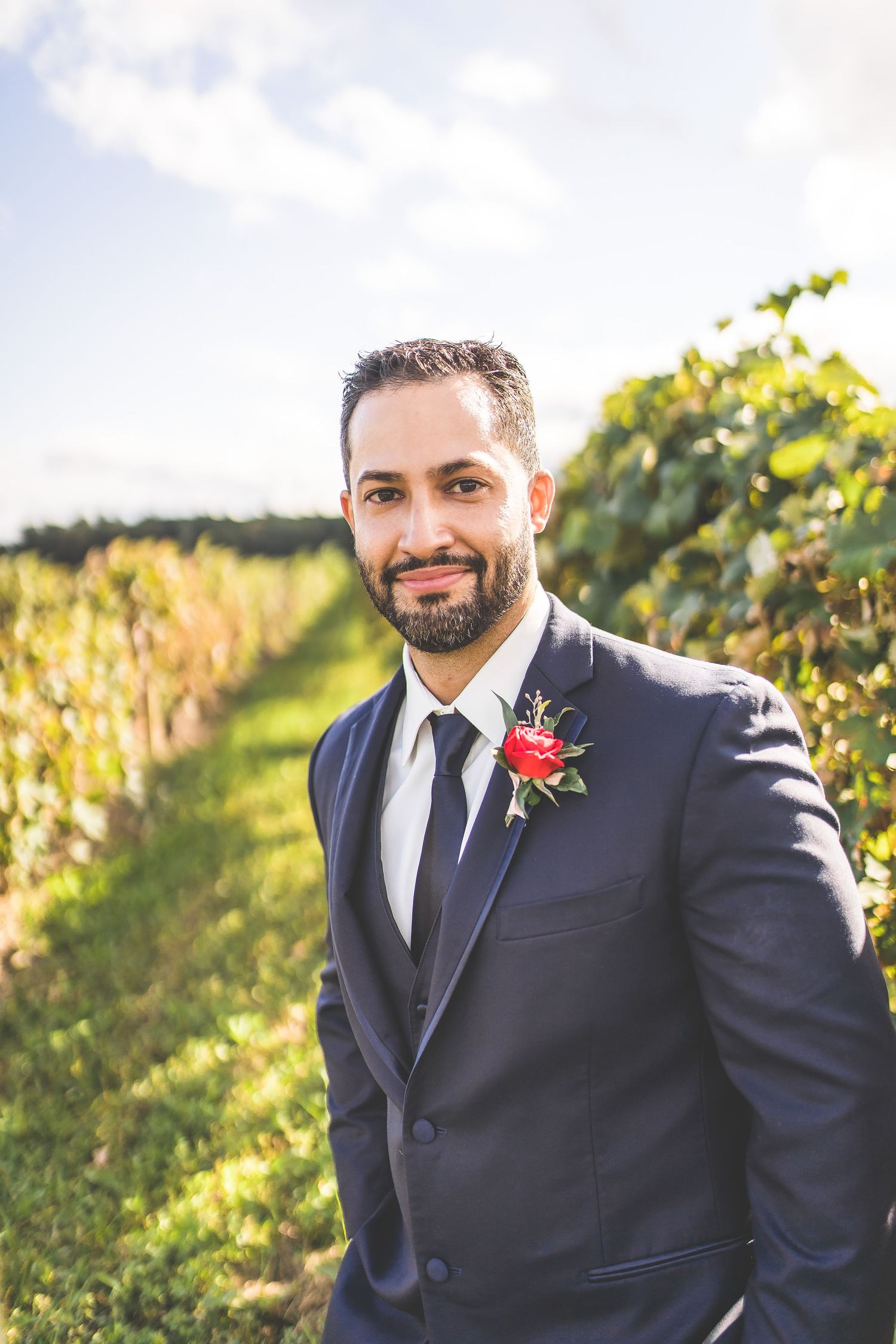 Groom in navy suit with red boutonniere poses in vineyard rows.