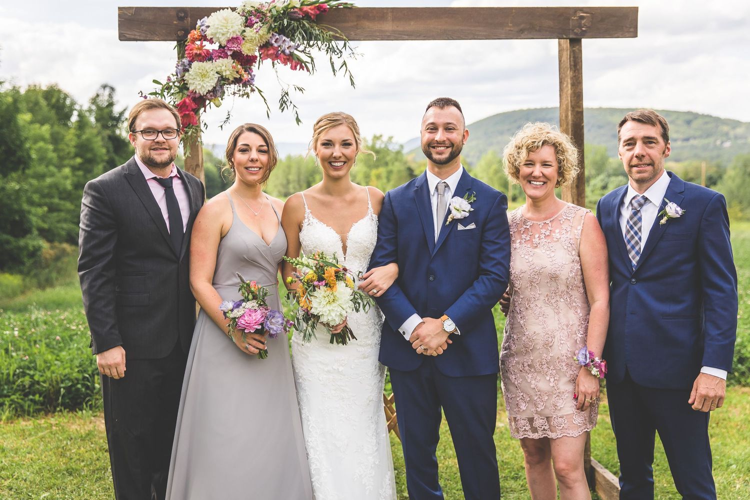 Wedding party poses under wooden arch with floral decorations in outdoor countryside setting.