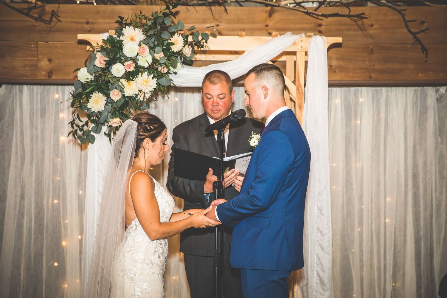 Wedding party dancing and celebrating at a rustic indoor reception venue with wooden beams and floral decorations.
