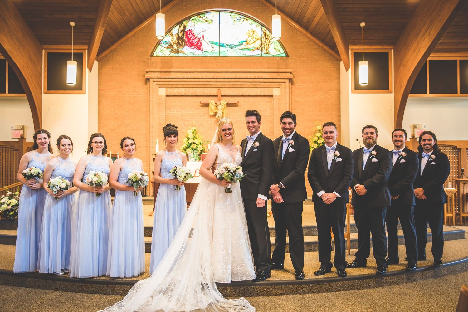 Wedding party poses in church sanctuary with bridesmaids in light blue dresses and groomsmen in dark suits.