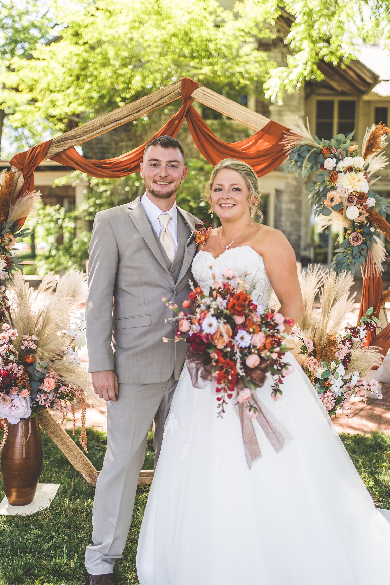A wedding couple poses under a rustic wooden arch decorated with orange fabric and floral arrangements in a garden setting.