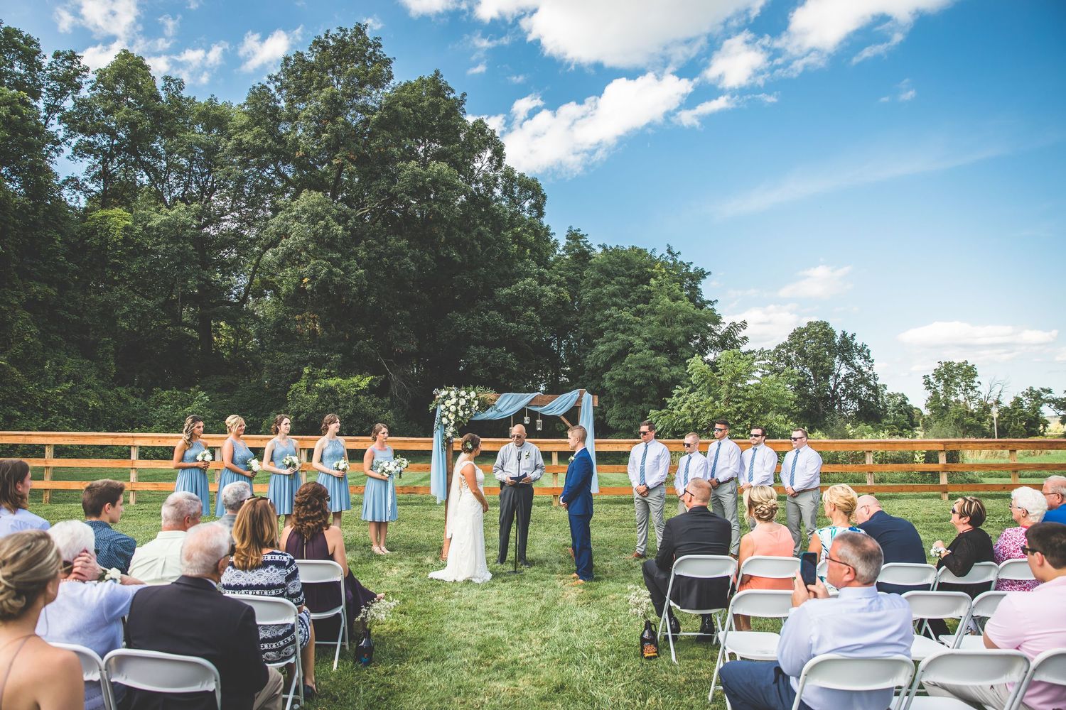 An outdoor wedding ceremony takes place in a grassy field with white chairs and wooden fencing.