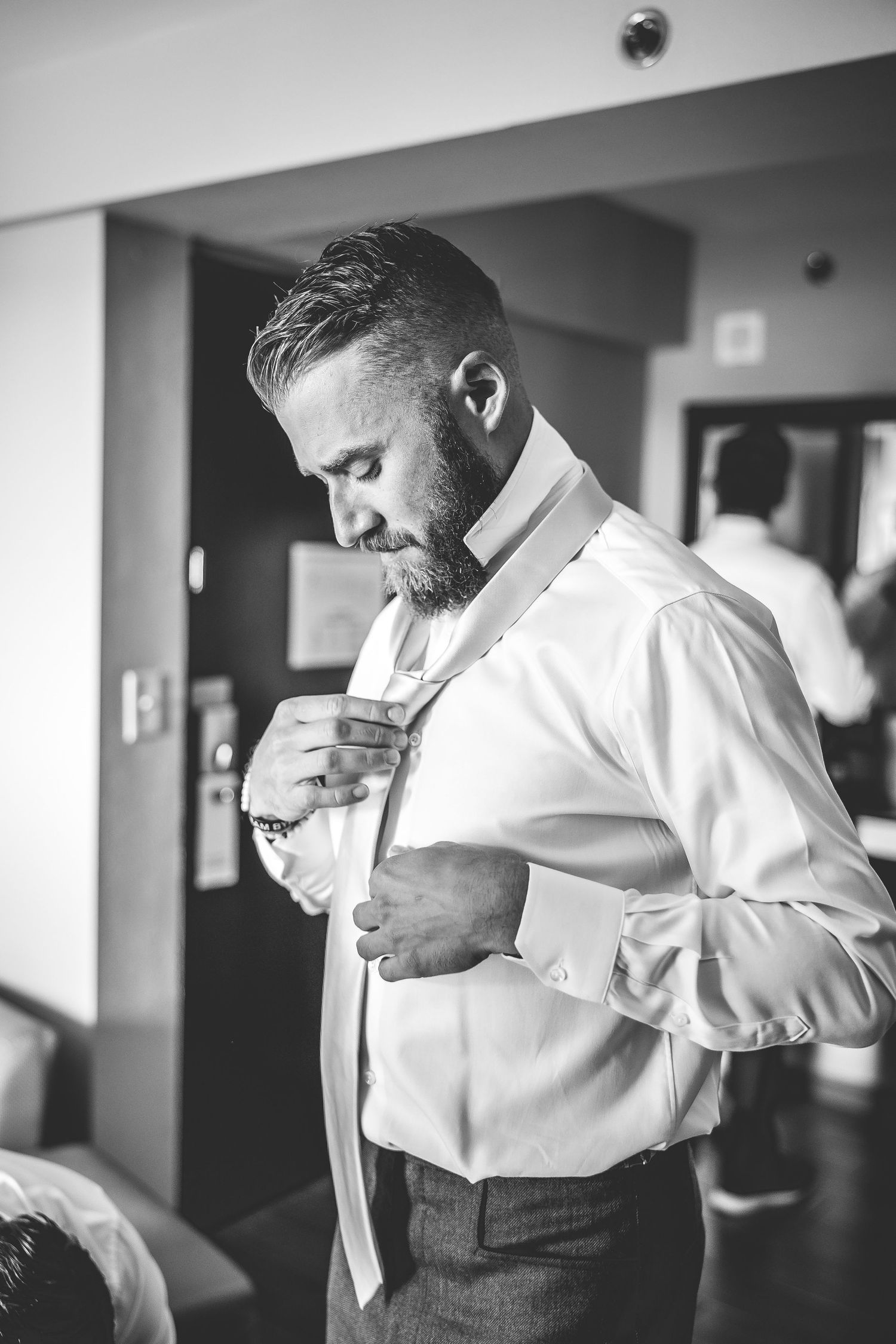 Black and white photo of groom adjusting tie while getting ready.