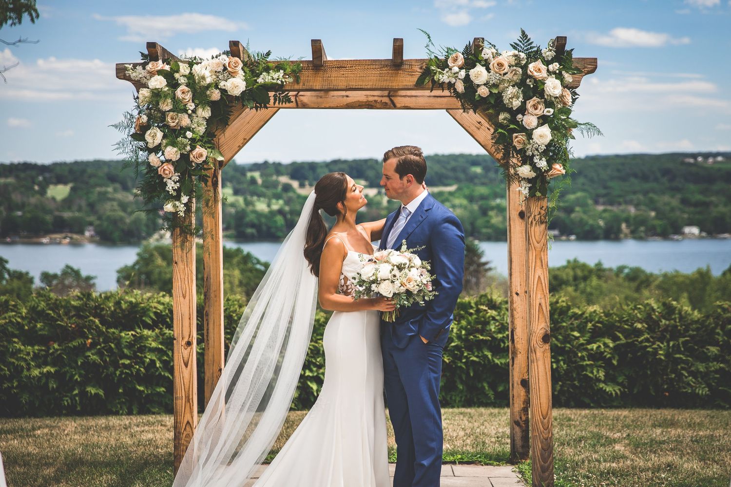 Wedding ceremony under a rustic wooden arch decorated with white floral arrangements against a blue sky.