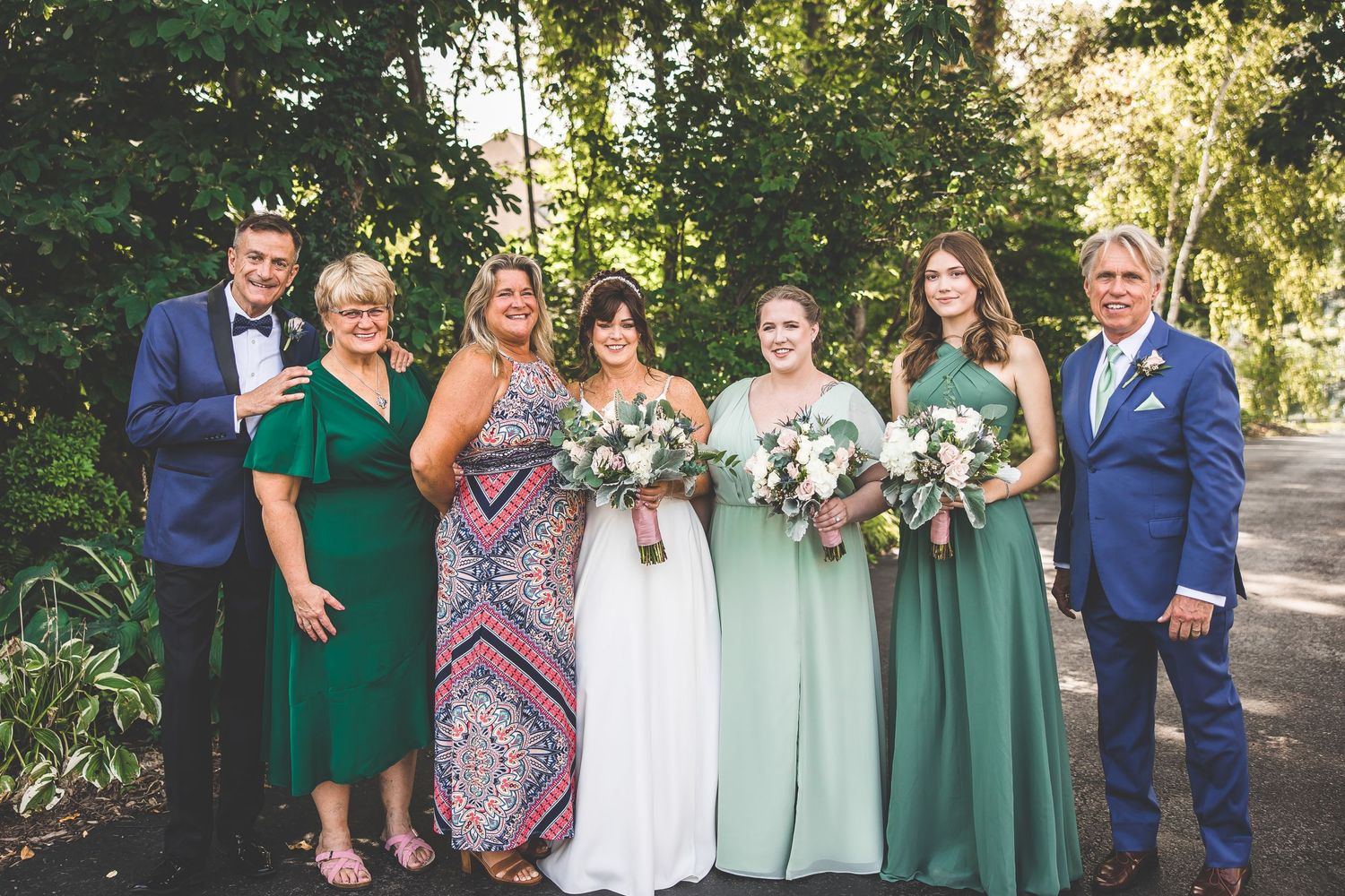 Wedding party poses in coordinating navy blue and sage green attire on a sunny outdoor patio.