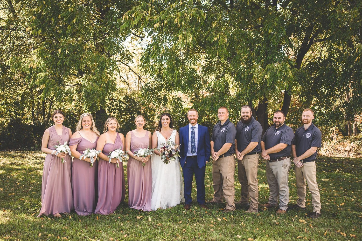 Wedding party in pink dresses and casual attire poses under trees for formal group photo.