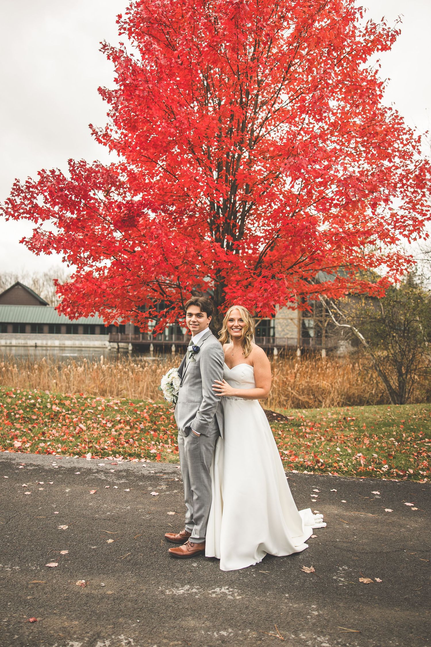 Wedding couple poses beneath stunning red maple trees on a fall day with scattered leaves.