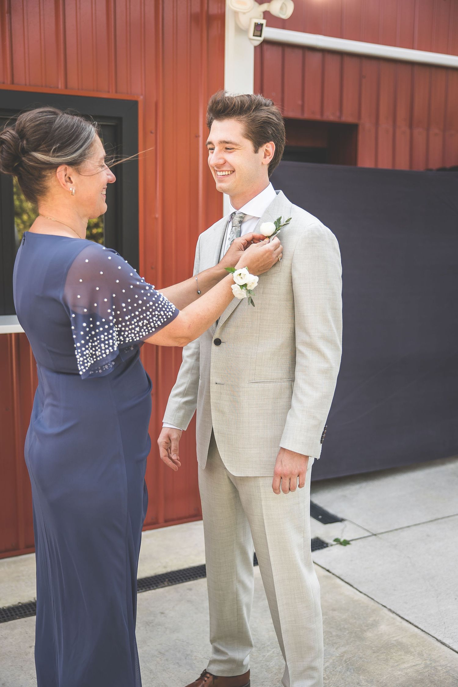 A touching moment as someone adjusts a light gray suit before the ceremony near a red barn.