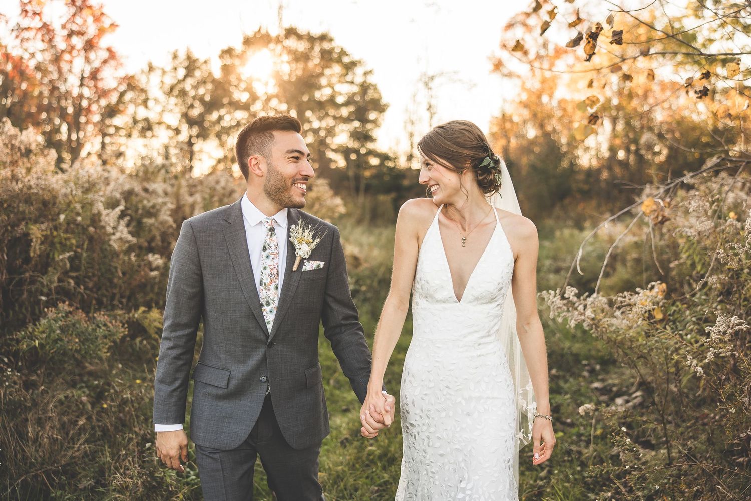 Bride and groom walk hand in hand through an autumn field at sunset.