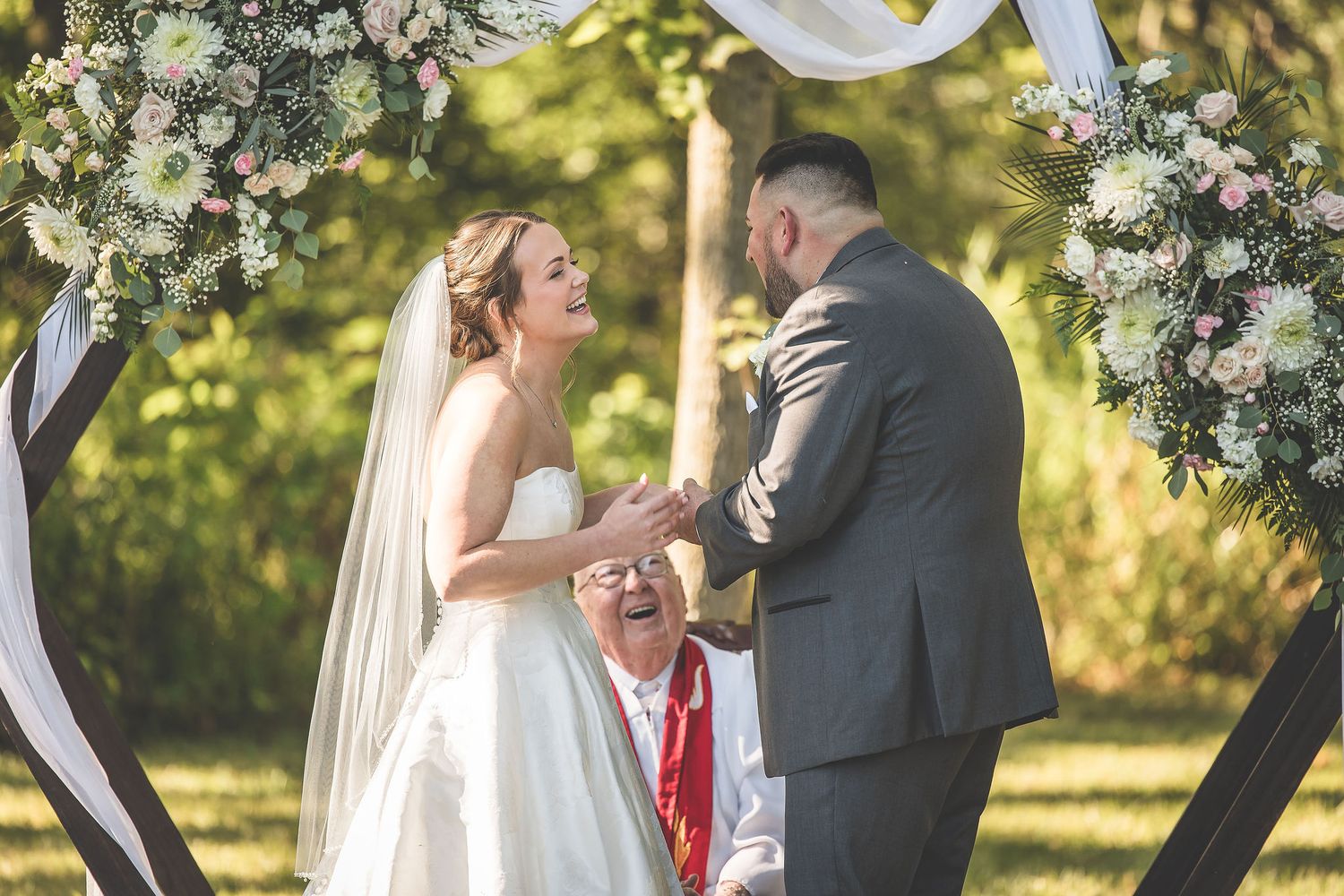 A romantic wedding ceremony moment under a decorated arch with white fabric and floral arrangements.