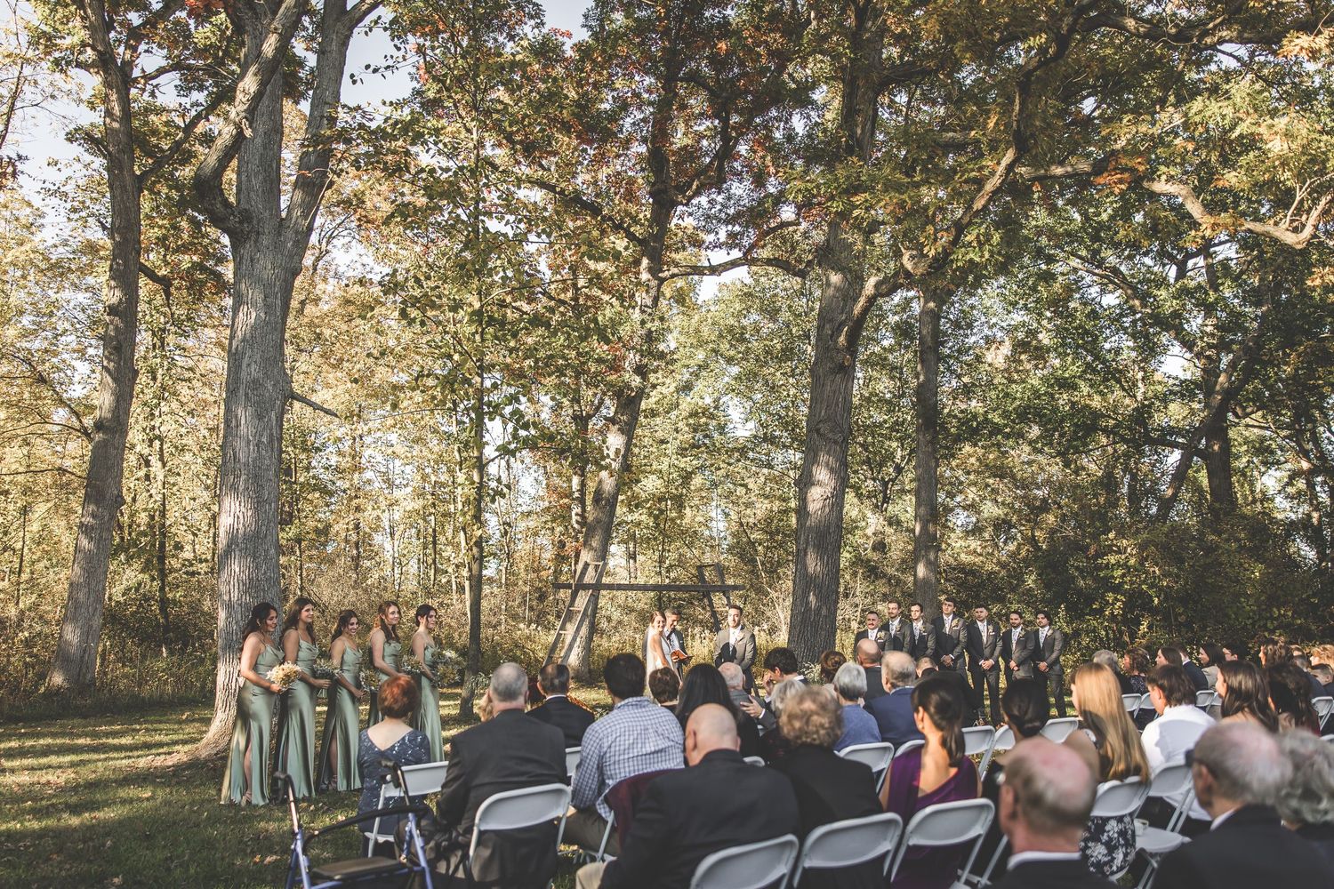 Wide view of outdoor wedding ceremony with guests seated among autumn trees.