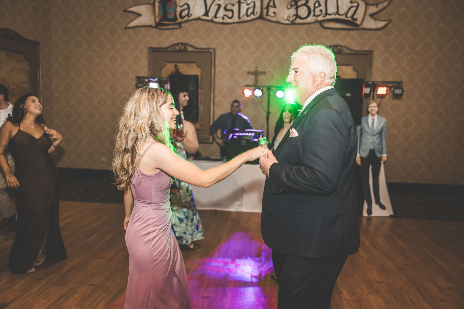 Wedding reception dance floor under a 'La Visible Bella' sign with colorful lighting.