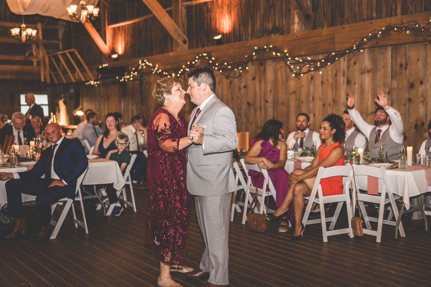 Guests observe couple dancing in rustic barn wedding reception with string lights and white chairs.