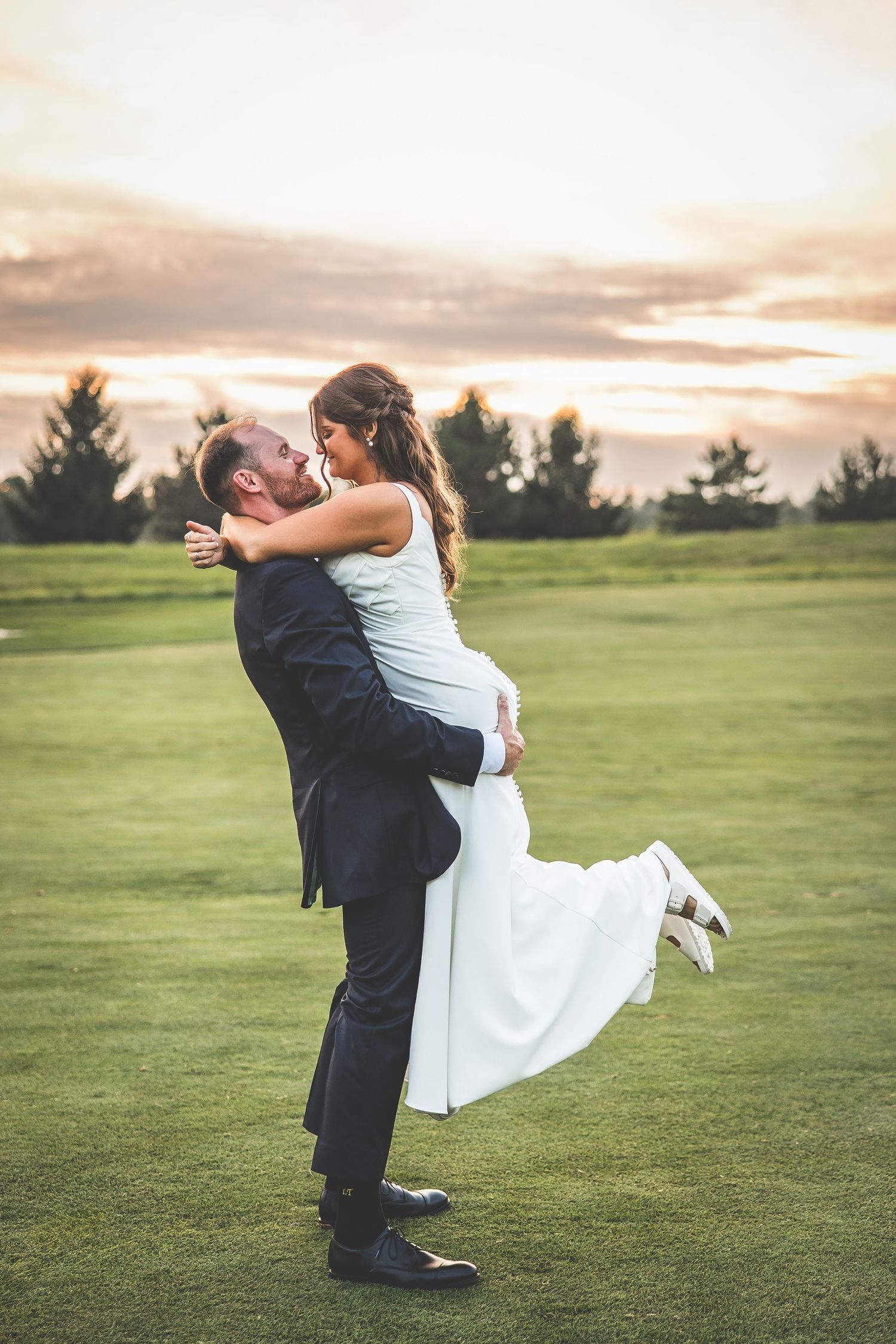 Playful wedding couple dancing on a lush green golf course at sunset.