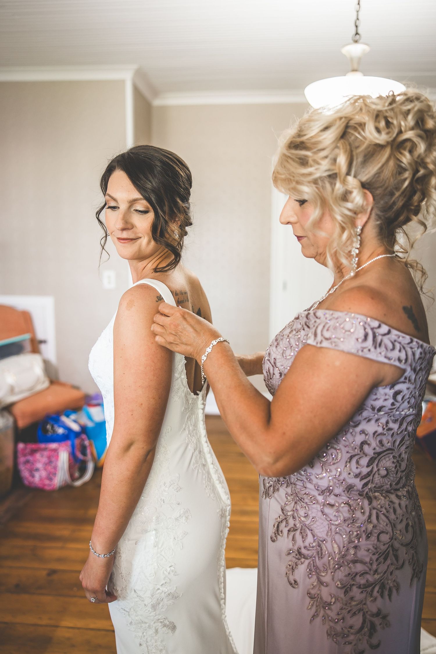 A bride getting help putting on her wedding dress from someone wearing a purple lace gown.