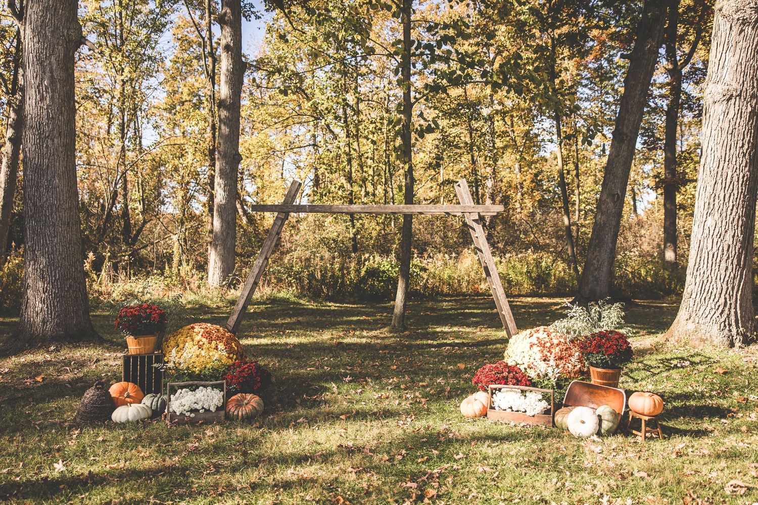 Rustic wooden arch decorated with fall flowers and pumpkins in a forest setting.