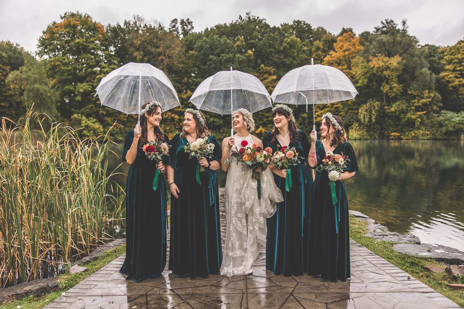 Bridal party with umbrellas poses by a lake wearing emerald velvet dresses and white wedding gown.